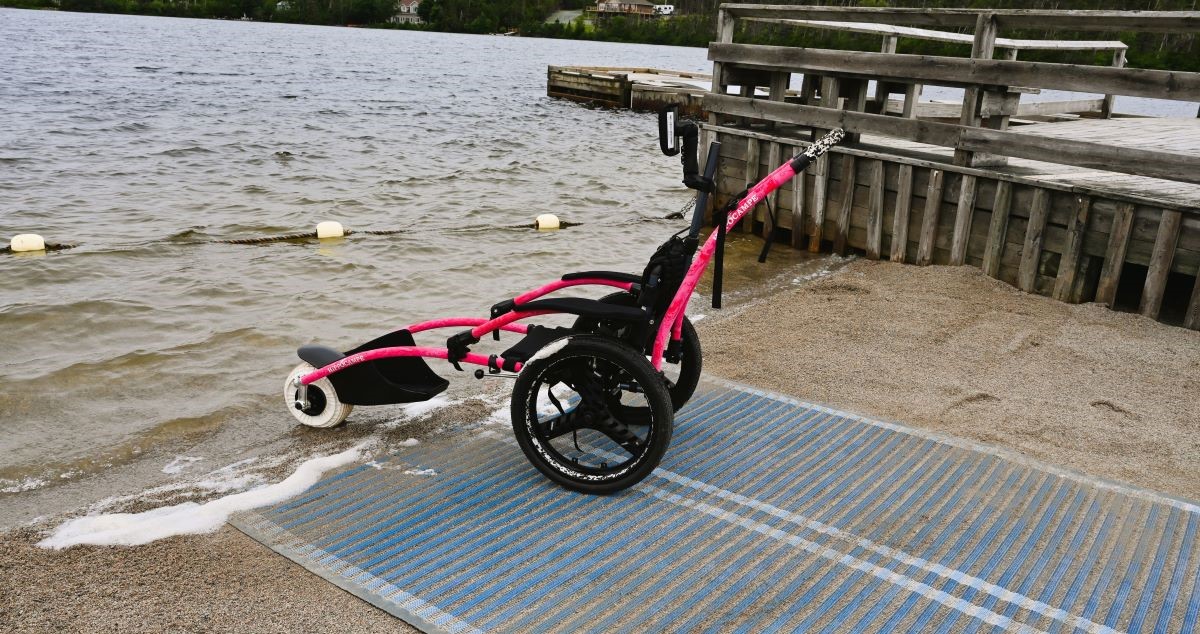  The image shows an all-terrain wheelchair positioned at the edge of a body of water at Rotary Sunshine Park. It is placed on a blue beach mat that leads from the sand into the water, ensuring accessibility for users. 