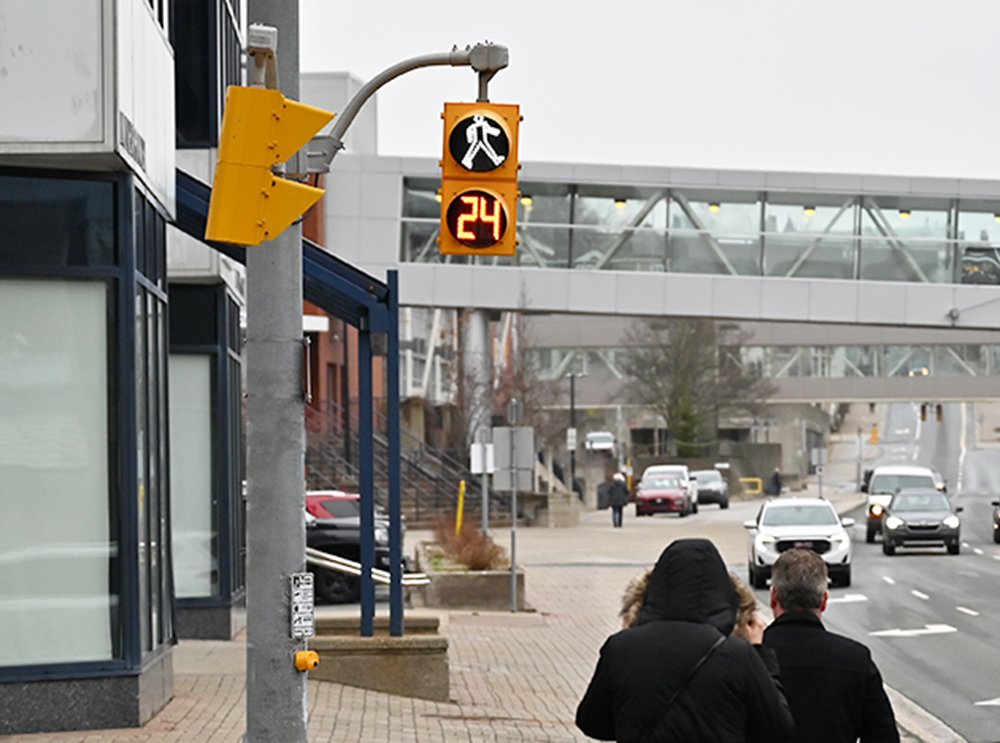 Two people wearing black are walking towards a grey building and a sidewalk. A pedestrian signal showing the 'walk' symbol with the number 24 is above the street crossing. 
