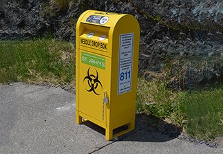A metal needle disposal box painted yellow about the size of a mailbox sitting on a pad of concrete